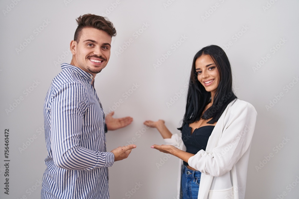 Young hispanic couple standing over white background inviting to enter smiling natural with open hand Young hispanic couple standing over white background inviting to enter smiling natural with open hand