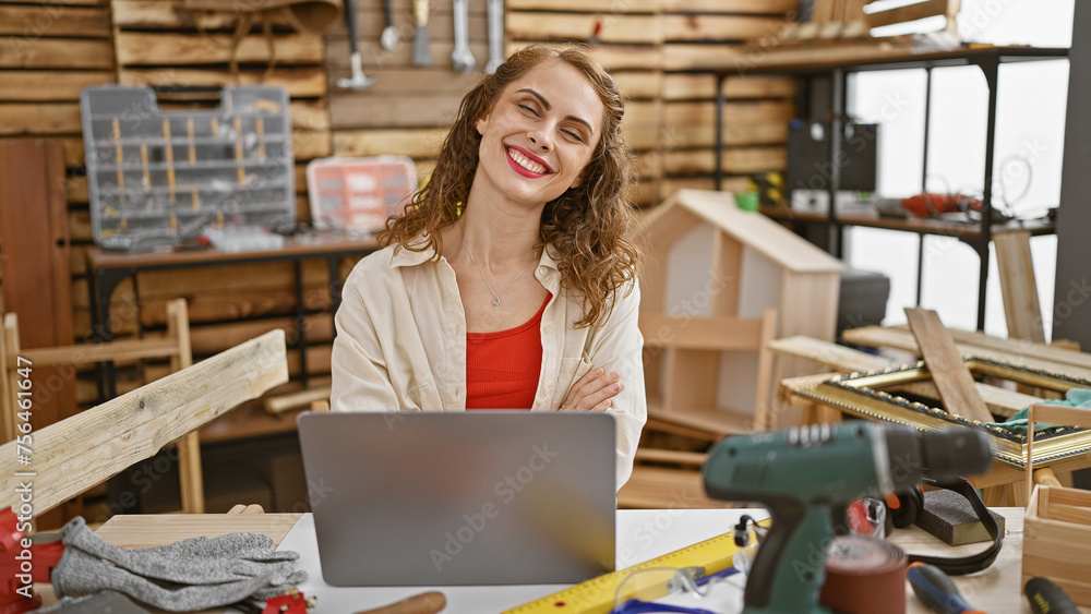 Smiling young woman carpenter engrossed in her online carpentry business, sitting at a table with laptop in her indoor workshop Smiling young woman carpenter engrossed in her online carpentry business, sitting at a table with laptop in her indoor workshop