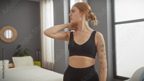 A young caucasian woman with tattoos stands in a modern bedroom, touching her neck in discomfort.