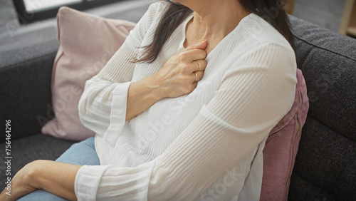 Middle-aged woman clutching chest in discomfort while seated on a sofa indoors, conveying potential health issues. photo
