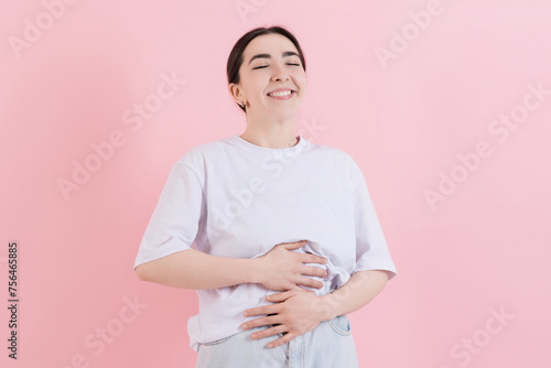 Caucasian young brunette woman in casual white t-shirt smiling and touching her stomach after a delicious healthy eating closing her eyes isolated on pink studio background.