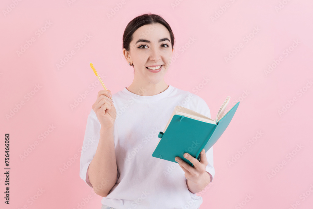 Attractive caucasian young brunette woman in casual white t-shirt has idea, writing notes in notebook, smiling and looking at the camera isolated on pink studio background. Attractive caucasian young brunette woman in casual white t-shirt has idea, writing notes in notebook, smiling and looking at the camera isolated on pink studio background.