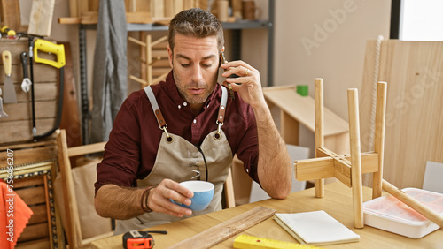 Hispanic man chatting on phone while holding a coffee cup in a carpentry workshop
