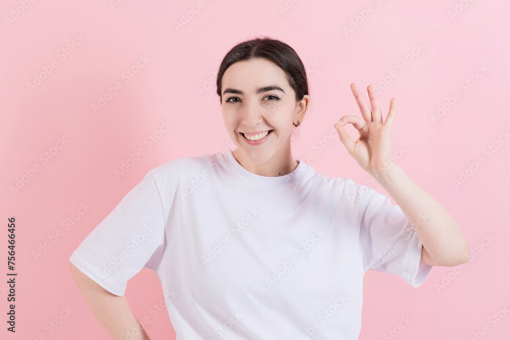 Attractive Caucasian young brunette woman in casual clothing looks at the camera, smiles and shows hand okay gesture isolated on pink studio background. Success, approval. Attractive Caucasian young brunette woman in casual clothing looks at the camera, smiles and shows hand okay gesture isolated on pink studio background. Success, approval.