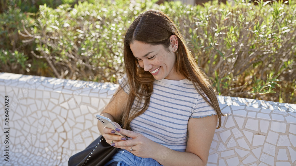 Smiling young woman with brunette hair using smartphone in sunlit park on a bench Smiling young woman with brunette hair using smartphone in sunlit park on a bench