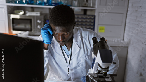 African male scientist feeling exhausted in the lab, leaning on microscope and computer