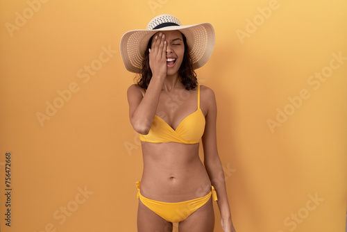 Young hispanic woman wearing bikini and summer hat covering one eye with hand  confident smile on face and surprise emotion.
