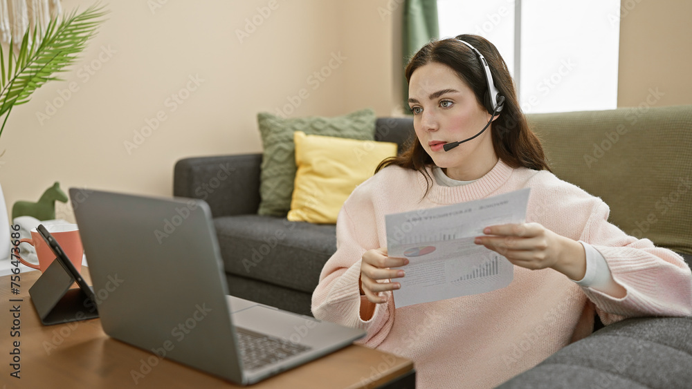 A focused woman wearing a headset reviews documents while working remotely on a laptop in a cozy living room. A focused woman wearing a headset reviews documents while working remotely on a laptop in a cozy living room.