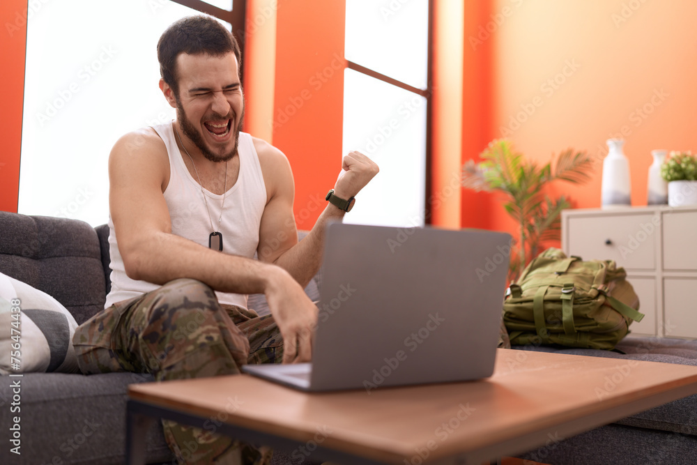 Young hispanic man wearing camouflage army uniform doing video call at home screaming proud, celebrating victory and success very excited with raised arm Young hispanic man wearing camouflage army uniform doing video call at home screaming proud, celebrating victory and success very excited with raised arm