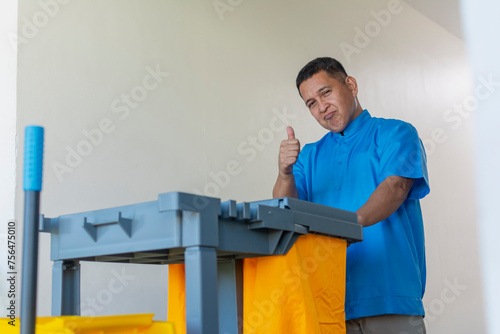 Middle-aged male janitor in blue uniform with janitorial cart showing thumbs up gesture in a bright hallway.
