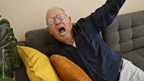 Caught in a cozy moment, portrait of a tired elder man with white hair and glasses waking up with a yawn, relaxing on a comfortable sofa in his home living room