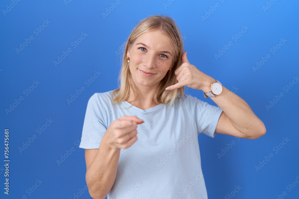 Young caucasian woman wearing casual blue t shirt smiling doing talking on the telephone gesture and pointing to you. call me. Young caucasian woman wearing casual blue t shirt smiling doing talking on the telephone gesture and pointing to you. call me.