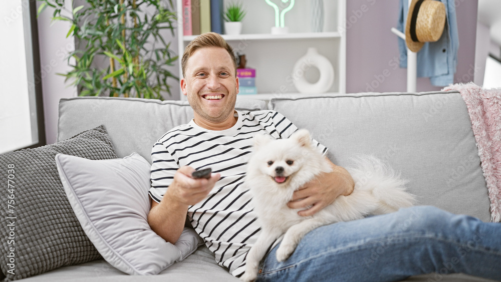 Handsome young caucasian man finding joy by relaxing with his happy pet dog, comfortably watching a movie on tv, smiling confidently while resting on the living room sofa at home.
