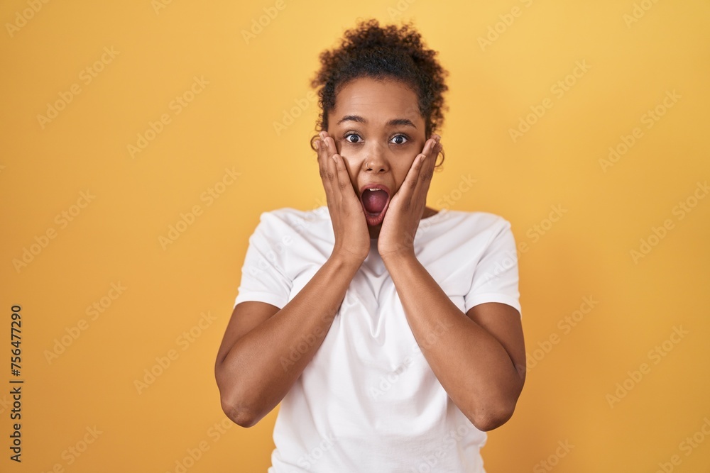 Young hispanic woman with curly hair standing over yellow background afraid and shocked, surprise and amazed expression with hands on face Young hispanic woman with curly hair standing over yellow background afraid and shocked, surprise and amazed expression with hands on face