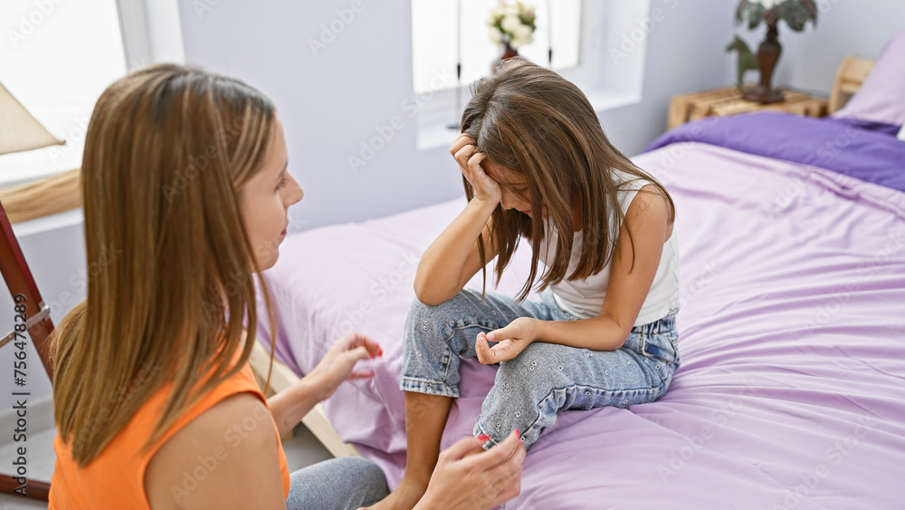 A caring woman comforts her distressed daughter sitting on a bed in a cozy home bedroom.
