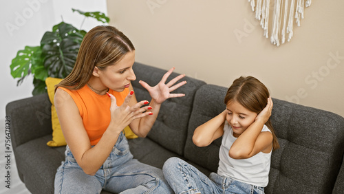 A frustrated woman gestures while speaking to a young girl covering her ears on a couch, implying a potential disagreement.