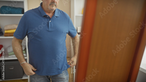 Mature man in blue shirt stands contemplatively in a room filled with shelves and clothing.