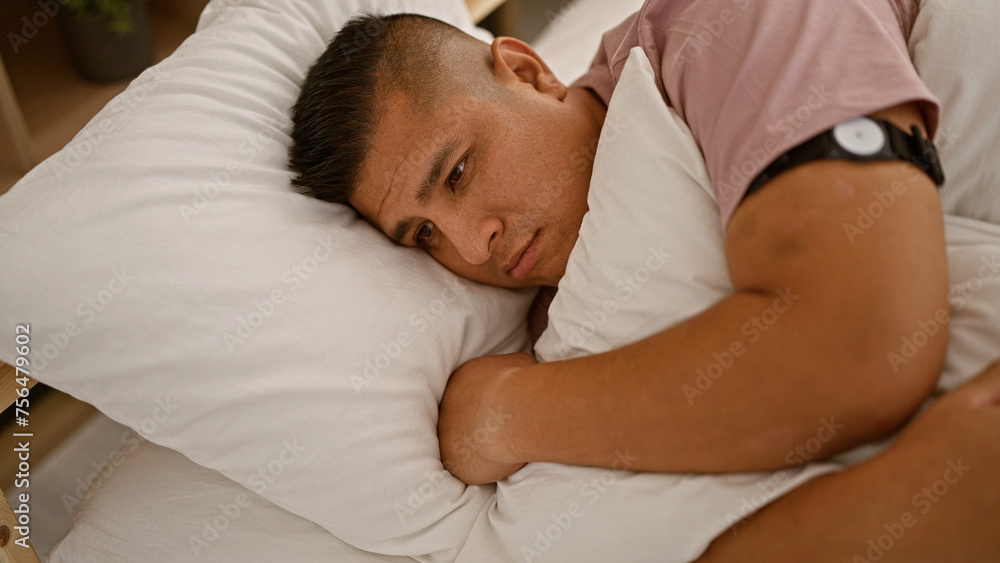 Sad young latin man hugging his pillow, laying in bed with a serious look, possibly facing morning glucose sensor levels problem in his bedroom