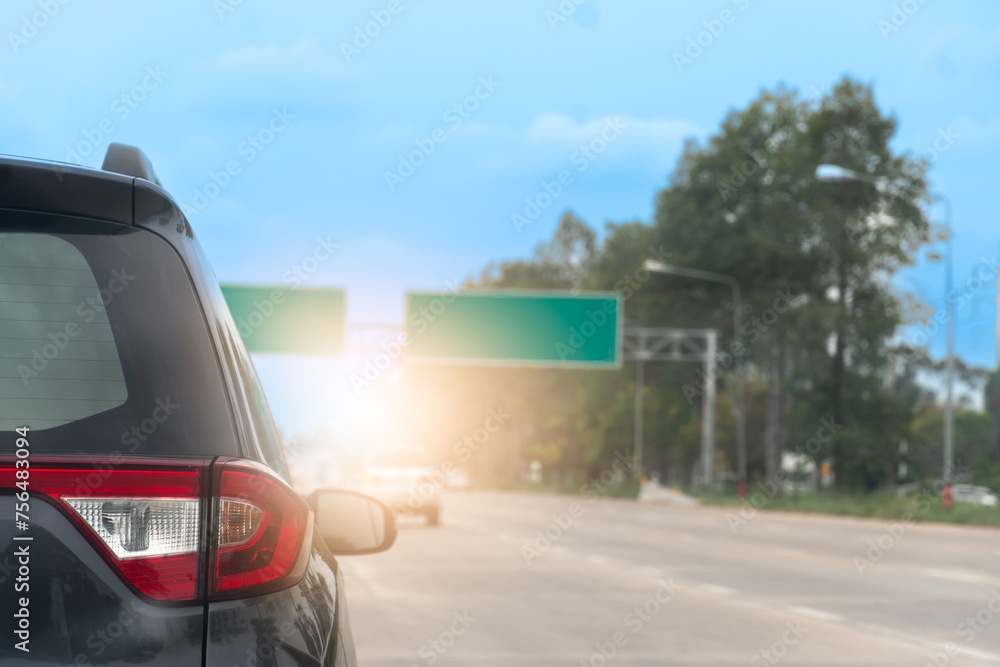 Rear side of a grey car on the road with other cars driving on the road. Sign borad green on top of road. Blurred background of trees under blue sky.