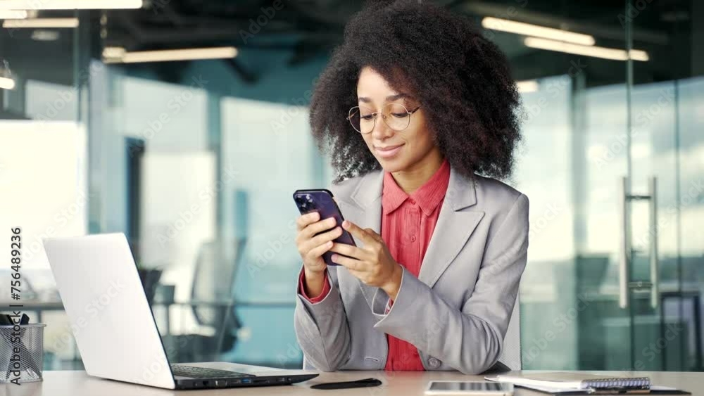Happy young african american businesswoman in a jacket is using phone sitting at workplace in business office. Black woman reading typing a message, chatting online and looking at the camera. Portrait Happy young african american businesswoman in a jacket is using phone sitting at workplace in business office. Black woman reading typing a message, chatting online and looking at the camera. Portrait