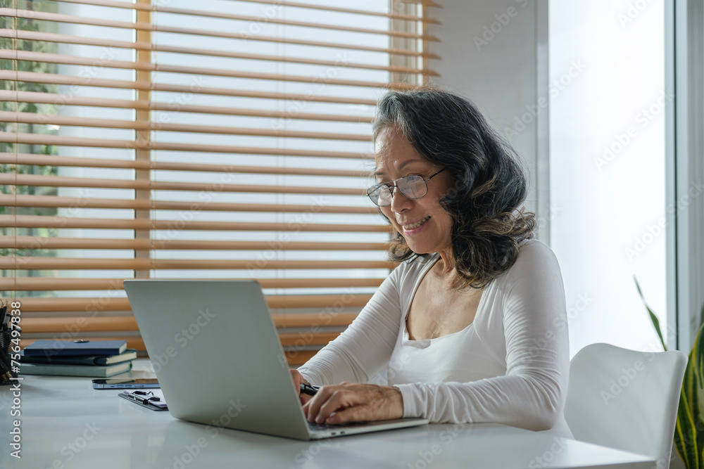 Beautiful mature woman in casual clothes sitting at table in home office and working on laptop computer. Beautiful mature woman in casual clothes sitting at table in home office and working on laptop computer.