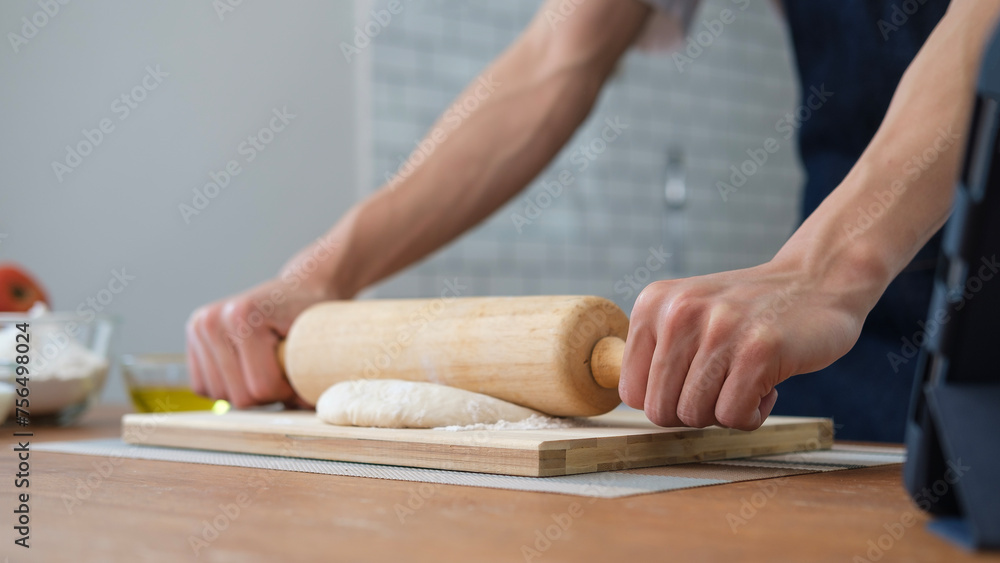 Unrecognizable young man using a rolling pin to roll over bread doughs on wooden table in kitchen.