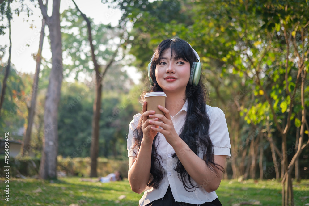 Obraz premium Satisfied young Asian woman listening to music with headphone and drinking coffee in the public park.