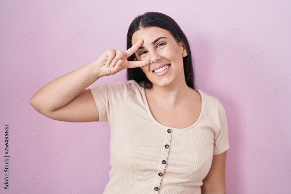 Young hispanic woman standing over pink background doing peace symbol with fingers over face, smiling cheerful showing victory Young hispanic woman standing over pink background doing peace symbol with fingers over face, smiling cheerful showing victory