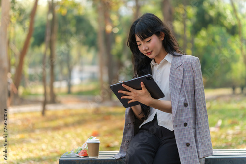 Charming young businesswoman using digital tablet while sitting on bench in the city park.