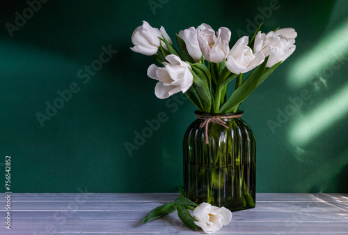 Bouquet of white tulips in green glass vase on green background with sunlight. Vase of flowers stands on wooden table. Concept: greeting card, spring holidays. Copy space