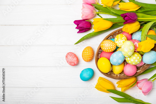 Easter basket filled with colorful eggs and a bouquet of tulips on a textured wooden table. Easter celebration concept. Colorful easter handmade decorated Easter eggs. Place for text. Copy space