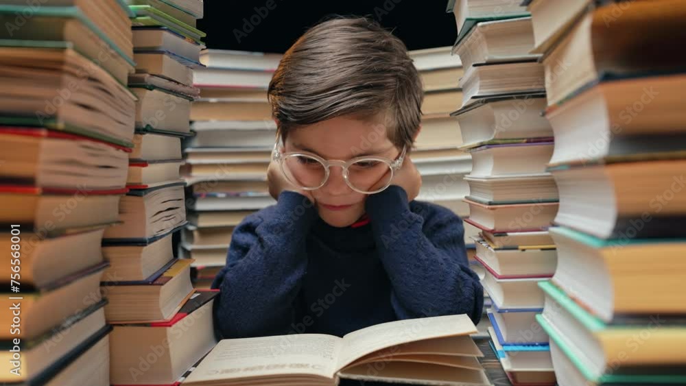 Cute little pupil boy in glasses reading interesting book in library between stacks of books literature. Education concept, prep or elementary school Cute little pupil boy in glasses reading interesting book in library between stacks of books literature. Education concept, prep or elementary school