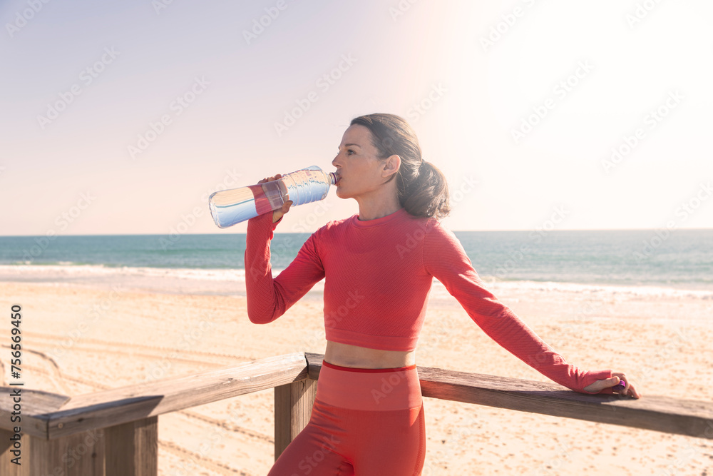 Αυτοκόλλητο sporty woman taking a break to drink and hydrate during exercise on the beach