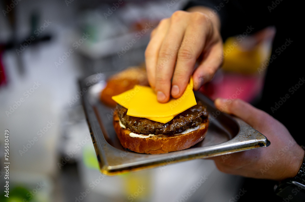 hand places a bright yellow slice of cheddar cheese onto a grilled burger patty, creating a cheeseburger. The burger sits on a toasted bun, and the background blurs, emphasizing the cheese placement.