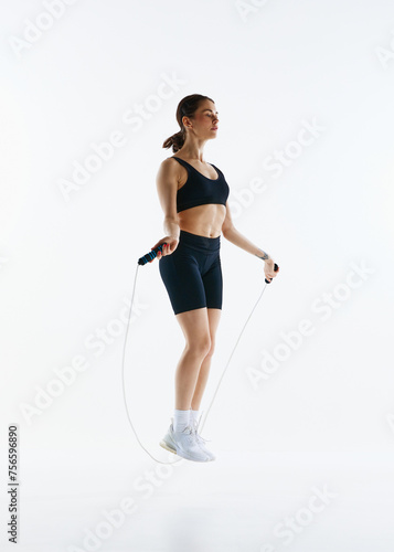 Full-length image of sportive slim young woman in sportswear training, doing exercises with jumping rope isolated over white studio background. Concept of sport, health and body care, fitness app