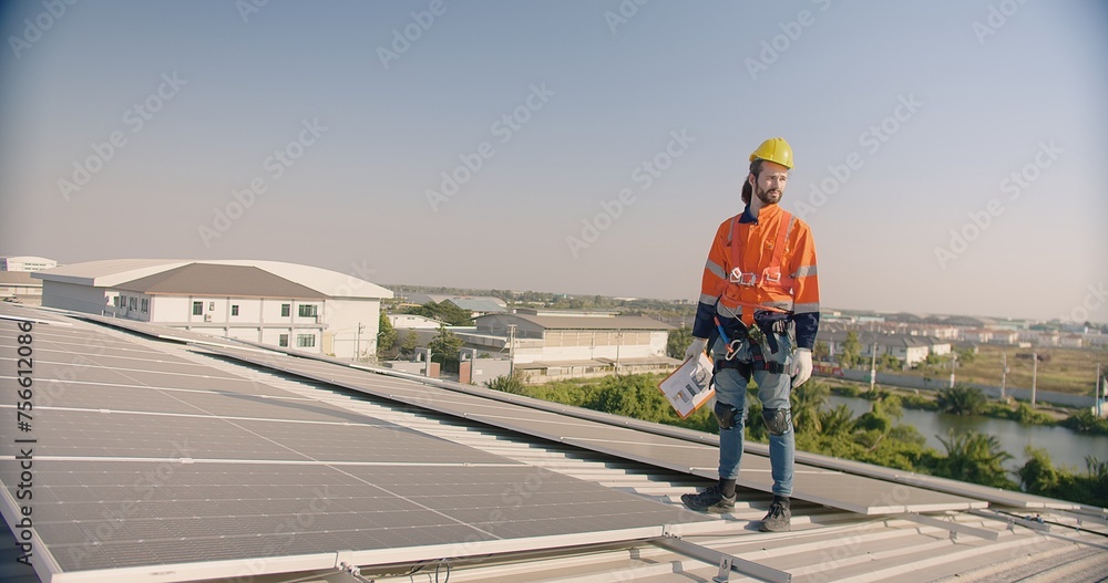 A solar panel technician engineer in safety harness and hard hat inspects a rooftop solar installation with a clipboard in hand against a clear sky and industrial landscape