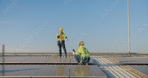 engineer Solar technicians in high-visibility jackets and safety helmets communicates using walkie-talkie while another sits with a tablet on a solar panel array on a rooftop solar farm under a clear