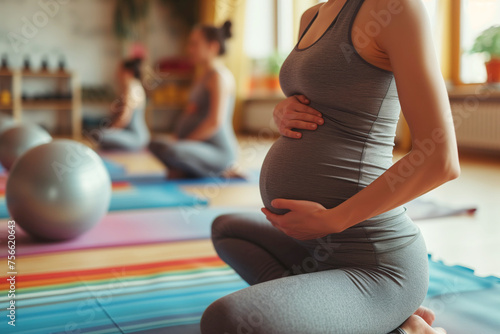 A pregnant woman in gym clothes sits on a yoga mat at a fitness gym and touches her belly