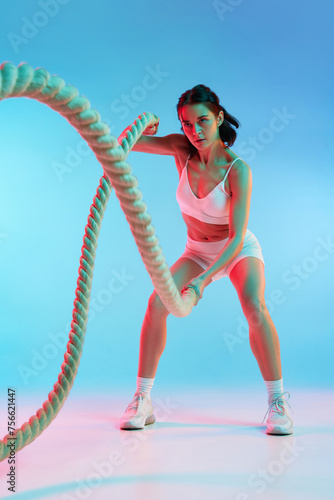 Sportive, slim young woman with strong muscular body training with ropes against blue studio background in neon light. Concept of sport, health and body care, fitness app, exercises templates