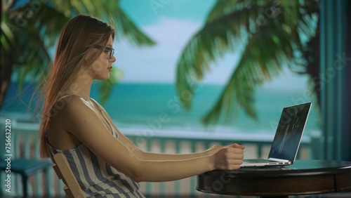 Female working from distance and travelling. Young woman freelancer working on laptop, sitting at the table on the tropical beach resort.