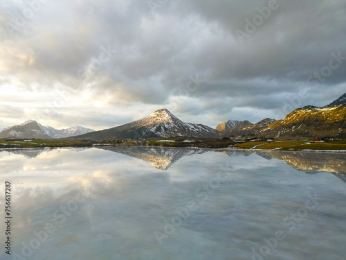 Panoramic view of beautiful white winter wonderland scenery with snowy mountain summits reflecting in crystal clear ice lake on a cold sunny day with sky and clouds. Norwegian fjords mountain.