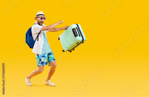 Happy funny male tourist going on summer vacation. Full body length joyful excited young man wearing holiday clothes, with travel suitcase and backpack walking on yellow orange color studio background photo