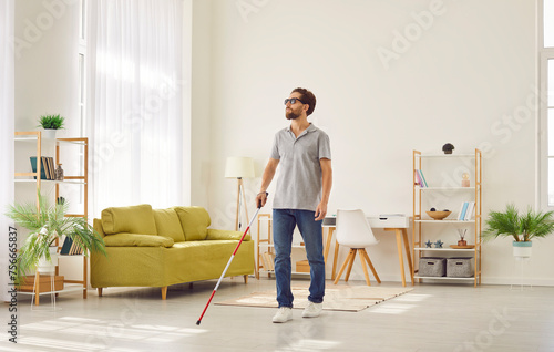 Full length portrait of blind young disabled man in casual clothes with cane walking in the living room at home. Male person with cane indoors. Blindness, disability and vision problems concept.