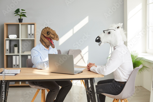 Strange people with funny, bizarre animal heads having a business meeting or a job interview. Two men wearing dinosaur and horse masks sitting at a table with a laptop in the office