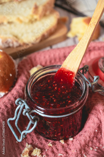 image of glass jar of raspberry or strawberry jam with a wooden knife. it is on a wooden table with slices of bread and a pink cloth.