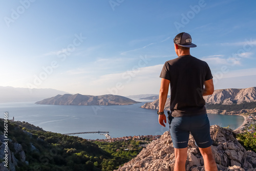 Hiker man on edge of rock cliff overlooking archipelago of Kvarner Bay in coastal town Baska, Krk Island, Primorje-Gorski Kotar, Croatia, Europe. Coastal hiking trail along coastline of Adriatic Sea