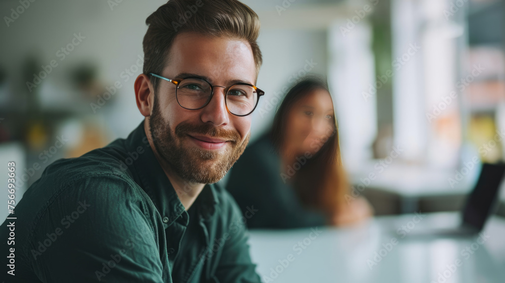 Young man with glasses in the foreground, smiling towards the camera, with coworkers in the background, in a meeting at office Young man with glasses in the foreground, smiling towards the camera, with coworkers in the background, in a meeting at office