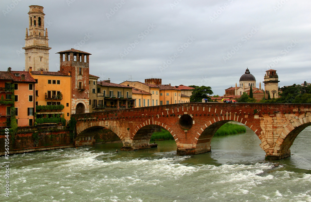 Cityscape view with the embankment, the Ponte Pietra brick arch bridge over the Adige river and the bell tower of the Cathedral against a stormy sky in Verona, Italy
