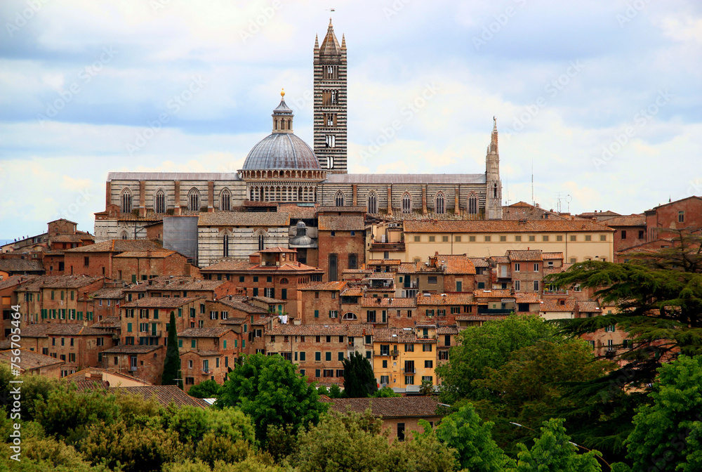 View of the historic part of the city of Siena with the Duomo di Siena and green trees and bushes in the foreground against a stormy sky in the Tuscany region of Italy