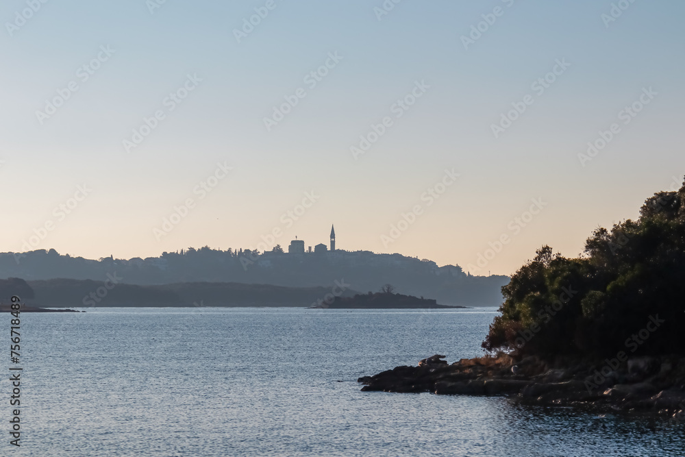 Distant silhouette view of coastal town Vrsar during dramatic sunset, Istria, Croatia. Calm sea surface reflects vibrant colors of sky. Vacation Adriatic Mediterranean Sea in summer. Church on hill